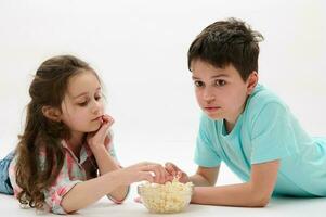 mooi gelukkig kinderen aan het liegen Aan buik, aan het eten popcorn Aan geïsoleerd wit achtergrond. kinderen kinderjaren levensstijl vrije tijd foto
