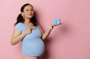 mooi latina zwanger vrouw richten vinger terzijde schattig blauw baby slofjes Aan haar hand, geïsoleerd Aan roze achtergrond. foto