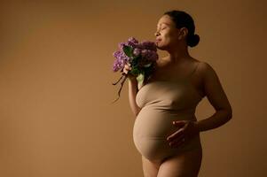 zwangerschap. moederschap. vruchtbaar. lichaam positiviteit. gravid vrouw in lingerie, met bundel van seringen Aan beige achtergrond foto