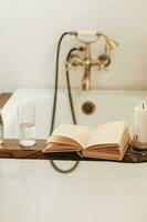 een boek, een fles van water, een glas en een kaars Aan een houten dienblad bovenstaand de badkamer, tegen de achtergrond van een antiek kraan foto