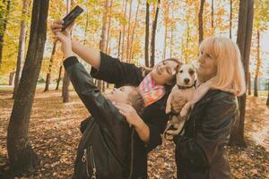 drie generaties van Dames en hond voelen pret kijken Bij camera poseren voor zelfportret afbeelding samen, grappig opgewonden kind, mam en grootmoeder hebben pret genieten weekend nemen selfie Aan apparaatje in herfst foto