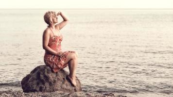blonde vrouw in paarse jurk zittend op de enorme rots naast de zee, kijkend naar de zonsondergang en wachtend op iets of iemand. foto