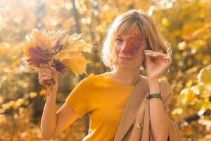 jong blond vrouw aan het bedekken een oog met rood esdoorn- blad. herfst en seizoen concept. buitenshuis vallen vrouw portret detailopname met gebladerte foto