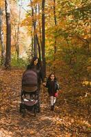 moeder en haar weinig dochter en een baby in kinderwagen Aan wandelen in herfst hout foto