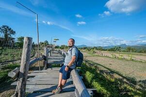 Aziatisch backpacker dik Mens met baard Aan de houten brug met blauw lucht in vakantie tijd foto