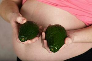 jong zwanger vrouw houdt twee avocado's dichtbij naar haar buik. foto