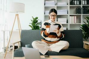 zelfverzekerd Aziatisch vrouw met een glimlach staand Holding kladblok en tablet in leven kamer Aan de sofa Bij huis foto