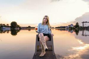 jong blond vrouw drinken bier zittend Aan de kade van een rivier- haven foto