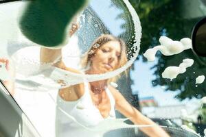 schattig midden- oud vrouw het wassen de auto glas met een veel van wasmiddel schuim foto