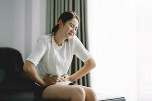 jong Aziatisch vrouw maag pijn zittend Aan bed Bij huis, Gezondheid probleem ontsteking in lichaam, periode fiets dag van maandelijks, menstruatie concept. foto