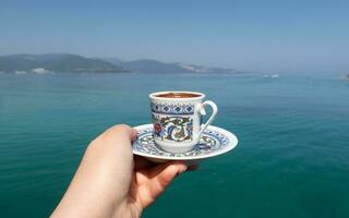 traditioneel Turks koffie kop met exotisch zee achtergrond, vrouw hand- Holding de kop Aan een zonnig zomer dag foto