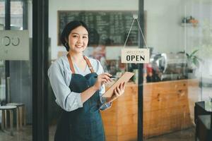 mooi jong barista vrouw in schort Holding bestellen papier en staand in voorkant van de deur van cafe met Open teken bord. bedrijf eigenaar opstarten concept. foto