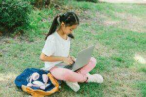 weinig meisje zittend Aan gras en gebruik makend van laptop in de park. onderwijs concept. foto