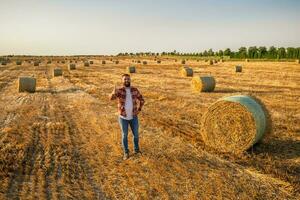 gelukkig boer is staand naast balen van hooi. hij is tevreden omdat van geslaagd oogsten. foto