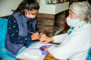 senior vrouw krijgen een manicure Bij huis gedurende covid 19 pandemisch vervelend gezicht masker foto