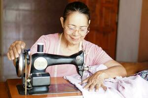 gelukkig senior Aziatisch vrouw kleermaker is naaien kleding door wijnoogst naaien machine Bij huis. concept, senior levensstijl, besteden vrij tijd naar reparatie of ontwerp kleren. hobby, niet alleen. foto