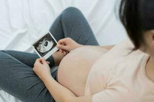 zwanger vrouw is op zoek Bij een echografie foto van foetus. moeder voorzichtig accenten de baby Aan maag. Vrolijk, familie, groei, zwangerschap, genot , bereiden pasgeboren, nemen zorg, gezondheidszorg, buik.