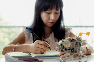 Aziatisch vrouw hand- schrijven Aan een notitieboekje met wazig geld en munten in een boodschappen doen kar foto