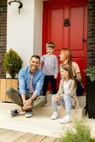 familie met een moeder, vader, zoon en dochter zittend buiten Aan de stappen van een voorkant veranda van een steen huis foto