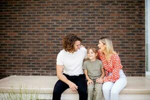 familie met een moeder, vader en dochter zittend buiten Aan de stappen van een voorkant veranda van een steen huis foto