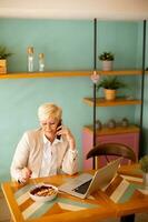 senior vrouw gebruik makend van mobiel telefoon terwijl werken Aan laptop en hebben gezond ontbijt in de cafe foto