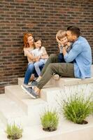 familie met een moeder, vader, zoon en dochter zittend buiten Aan de stappen van een voorkant veranda van een steen huis foto