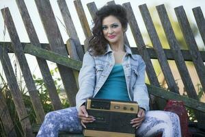 een mooi land meisje in helder kleren zit Aan een houten bank met een cassette recorder. vrouw in de stijl van de jaren 90. foto