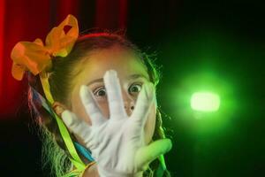 de weinig actrice meisje gedekt haar gezicht met een wit handschoen en looks door haar vingers. stadium licht en kind acteur. foto