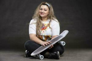 mooi dik vrouw met een skateboard Aan een grijs achtergrond looks Bij de camera en glimlacht. foto