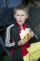 een grappig weinig jongen is hongerig en is aan het eten een groot broodje. foto