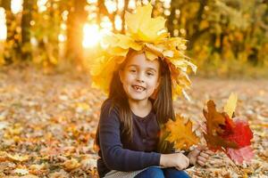 portret van glimlachen kind met krans van bladeren Aan hoofd. achtergrond van zonnig herfst park. foto