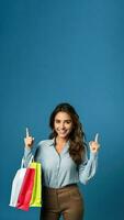 glimlachen jong brunette vrouw Holding boodschappen doen Tassen met aankopen na boodschappen doen punt vinger terzijde geïsoleerd Aan blauw achtergrond foto