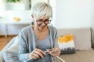 ouderen vrouw in bril zitten Aan bankstel Bij huis glimlach Holding breiwerk naalden en garen breit kleren voor geliefde degenen, favoriete werkzaamheid en tijdverdrijf, gepensioneerd rustig zorgeloos leven concept foto