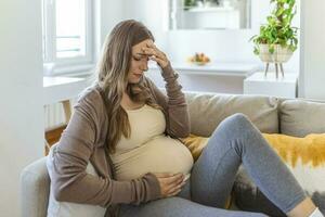 zwanger jong volwassen vrouw resting Aan sofa Bij huis, gevoel onwel. jong zwanger vrouw heeft geleden van hoofdpijn zittend Aan de bank. zwangerschap symptomen foto