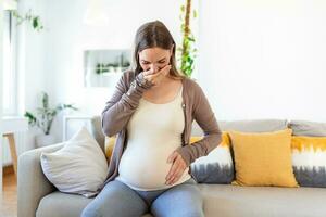 jong zwanger vrouw lijden van toxicose Bij huis. moederschap dame hebben ochtend- ziekte aan het bedekken mond met handen terwijl zittend Aan bankstel in leven kamer. mam verwachten ongeboren baby foto