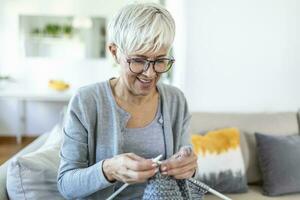 ouderen vrouw in bril zitten Aan bankstel Bij huis glimlach Holding breiwerk naalden en garen breit kleren voor geliefde degenen, favoriete werkzaamheid en tijdverdrijf, gepensioneerd rustig zorgeloos leven concept foto