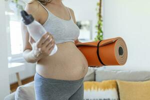 bijgesneden schot middenstuk van zwanger vrouw Holding een yoga mat en een herbruikbaar water fles, krijgen klaar naar oefening Bij huis. welzijn. blijven fit en gezond gedurende zwangerschap foto