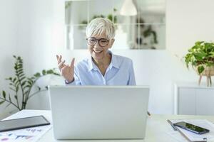 vrolijk volwassen vrouw hebben een video telefoontje van huis, online bedrijf concept. senior vrouw uitleggen met haar handen. bedrijf, zakenvrouw hebben video conferentie door laptop computer Bij kantoor foto