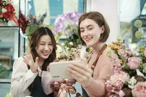 e-commerce bedrijf partner, twee jong vrouw bloemist arbeiders tonen bloemen regeling, online leven streamen, en selfies met smartphone toepassing in helder bloem winkel, mooi bloesems op te slaan. foto