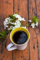 een kop van koffie Aan een donker, versleten rustiek houten tafel. de samenstelling is versierd met een takje met wit bloemen. kers boom bloemen. selectief focus. foto