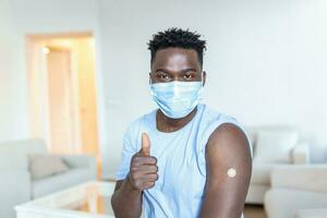 gevaccineerd Mens met beschermend gezicht masker op zoek Bij arm met injectie gips, portret van gevaccineerd Afrikaanse Mens tonen zijn arm foto