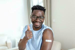 portret van een Mens glimlachen na krijgen een vaccin. Afrikaanse Mens Holding naar beneden zijn overhemd mouw en tonen zijn arm met verband na ontvangen vaccinatie. foto