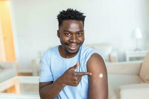 portret van een Mens glimlachen na krijgen een vaccin. Afrikaanse Mens Holding naar beneden zijn overhemd mouw en tonen zijn arm met verband na ontvangen vaccinatie. foto