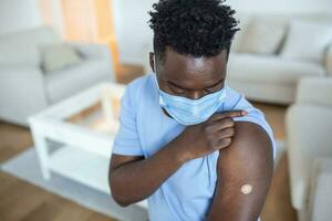 gevaccineerd Mens met beschermend gezicht masker op zoek Bij arm met injectie gips, portret van gevaccineerd Afrikaanse Mens tonen zijn arm foto