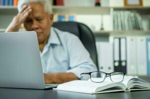 gelukkig senior Aziatisch gebruik makend van de laptop voor berekenen en maken een Notitie van dagelijks uitgaven. concept van levensstijl, aan het leren, en financiën bedrijf foto