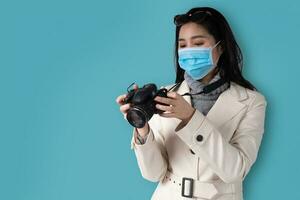 jong mooi Aziatisch vrouw Holding een camera en vervelend een medisch gezicht masker Aan een blauw geïsoleerd achtergrond, concept van reiziger toerist en vakantie. foto
