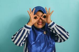 middelbare leeftijd Aziatisch vrouw in blauw hijaab, gestreept shirt, gebaren OK Leuk vinden verrekijker, ogen gluren door vingers. geïsoleerd Aan blauw achtergrond. foto