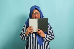 geërgerd Aziatisch middelbare leeftijd vrouw, vervelend een blauw hijaab, bril, en een gestreept shirt, houdt een Open boek en looks weg terwijl staand tegen een blauw achtergrond. foto