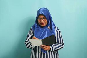 opgewonden Aziatisch middelbare leeftijd vrouw, vervelend een blauw hijab en een gestreept shirt, schrijft in een notitieboekje met enthousiasme terwijl staand tegen een blauw achtergrond. foto