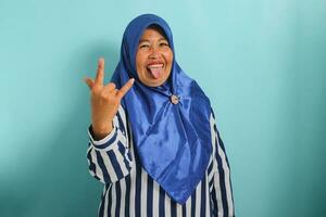 een vrolijk middelbare leeftijd Aziatisch vrouw in een blauw hijab en gestreept overhemd maakt de rots teken, gebaren met hand, tonen haar haar tong en liefde voor zwaar metaal, terwijl geïsoleerd Aan een blauw achtergrond. foto